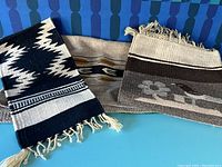 Three small to medium sized wool rugs with Native American and Southwestern patterns arranged overlapping on a blue surface against a blue patterned background. Rugs include a black and white zigzag geometric pattern, a gray gradient rug with bird and flower designs, and an earth tone patterned rug with horizontal stripes.