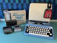 Photo showing typewriter in case, Sony Walkman players, Remo drum pad box, Garmin radio box, Winston headphones and additional small silver item on blue background