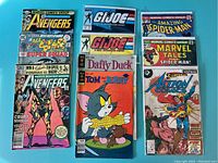 Photo showing a variety of vintage comic books arranged in two vertical stacks, featuring Avengers, G.I. Joe, Tom and Jerry, Daffy Duck, Spider-Man, Marvel Tales, Action Comics with Superman, Whitman Avengers, and All Star Comics Super Squad covers.