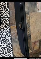 Photo showing black retractable cargo cover with handle, placed on floor tiles and near a patterned rug.