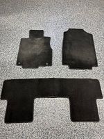 Three black car mats laid out on a surface: driver mat with mounting holes, passenger mat with irregular shape, and a wide second row mat.