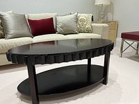 Photo of the dark wood oval coffee table featuring scalloped edging, showing front and side angled view with sofa and pillows in background.