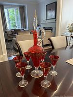 Full set of one red and gold decorative decanter with six matching glasses arranged on dark wood table in well-lit room.