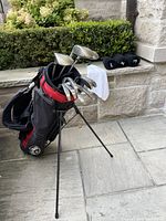 Full view of the golf bag standing with clubs inside, including woods and irons visible, with three black club head covers laid on the stone ledge.