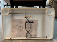 View of the underside of the tray table showing metal folding leg mechanism and instruction sheet labeled 'BAGN' in original packaging plastic wrap.