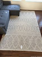 Full view of the beige woven area rug placed on hardwood floor next to grey sectional sofa.