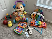 Overview of all toys included in the lot placed on carpeted floor near window showing the Fisher-Price Puppy Walker, tiger xylophone, piano keyboard, rattles, plush dog, and other baby toys.