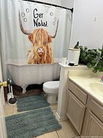 Shower curtain with highland cow and 'Get Naked' text, two teal bath mats on floor, plunger/toilet brush set on floor near toilet, white waste basket with bamboo lid on counter with faux plant.