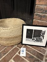 Photo of the round woven ottoman and framed black and white cat print showing front view of art and poof near a fireplace.
