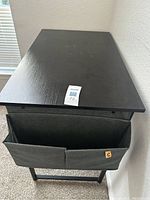 Top and side view of the black writing desk showing the wood grain tabletop and attached fabric storage pockets on one side.