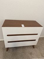 Front view of the modern two-drawer chest showing white fronts and wood grain top and legs.