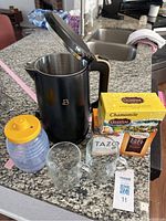Electric water boiler kettle with lid open on granite countertop with honey container, three tea boxes, two glass mugs, and MaxSold lot tag.