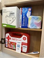 Cabinet shelf containing one large red all-purpose first aid kit box (160 pieces), one small multi-color translucent first aid case, two boxes of disposable face masks (KN95 and CVS Health brand).