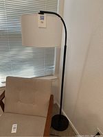 Full view of black metal floor lamp with curved arm and white drum shade next to a chair and window blinds.
