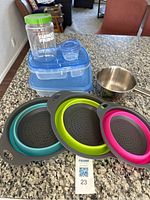 Photo showing three collapsible strainers in blue, lime green, and pink arranged next to two rectangular blue plastic food storage containers with lids, a small stainless steel saucepan, a labeled clear plastic jar, and a small plastic container with lid on a granite countertop.