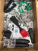 Drawer containing kitchen food prep utensils including can openers, baster brushes, peelers, small strainer, and a cookie cutter shaped like a winter sweater