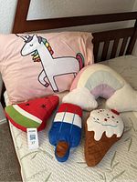 Photo showing unicorn pillowcase on pillow, pastel rainbow cloud pillow, and food-themed plush pillows (watermelon, popsicle, ice cream cone) arranged on a mattress.