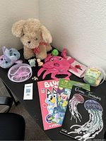 Photo showing tan plush puppy dog, pink reversible octopus, two squishy animal toys, three coloring books, and a clear plastic container with green lid containing small toys on a black table.