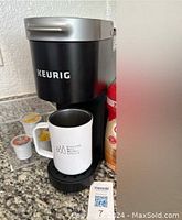 Black Keurig coffee maker on granite counter with white insulated mug placed on drip tray and three coffee pods alongside.
