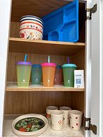 Wood cabinet shelf with blue 5-section tray, 5 plastic multi-colored cups with lids (2 with straws), and 4 ceramic cups with floral and letter R prints.