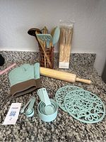 Overview of kitchen items on granite counter including turquoise oven mitts, trivets, measuring cups and spoons, wooden rolling pin, wooden skewers, and stirring utensils in copper holder.