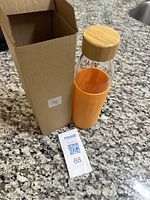 Glass tumbler with orange silicone grip and wooden cap shown next to its original brown box on a granite countertop.