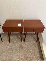 Pair of matching wooden nightstands side by side on carpeted floor with a white wall background. Both have a single drawer and modern tapered legs.