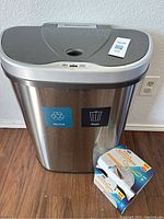 Front view of the Ninestars automatic trash can with recycle and trash labels and box of kitchen trash bags next to it.