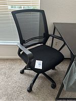 Front side view of black mesh office chair positioned next to desk and window, showing seat cushion and armrests.