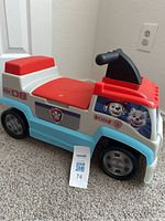 Side view of the ride-on toy showing Paw Patrol character decals, seating area with red seat, black handlebar steering, and four wheels.