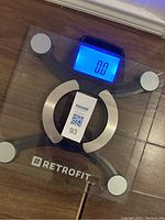 Top view of the digital bathroom scale powered on, showing 0.0 on the blue backlit display, placed on a wooden floor.