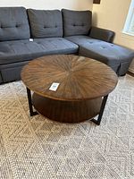 Round coffee table in living room setting, showing top sunburst wood grain pattern and lower shelf.