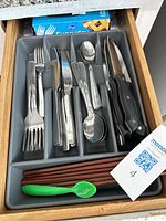 Drawer organizer with 4 small forks, 4 large forks, 8 butter knives, 5 small spoons, 6 large spoons, 8 steak knives with black handles, 1 green plastic baby spoon, 7 brown wooden chopsticks, and box of plastic disposable forks.