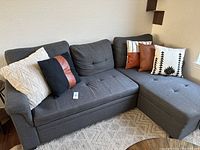 Photo showing five throw pillows arranged on a dark gray sectional sofa, highlighting colors and styles including cream textured, black with brown stripe, brown faux leather, white with navy stripes, and white with black southwestern pattern.