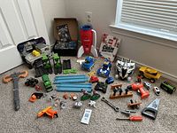 Wide shot showing a variety of toy vehicles including military trucks, cars, toy weapons, blue track pieces, and toy tools.