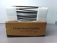 5-light drum chandelier in its original box with foam packaging, showing the black metal frame wrapped with horizontal metal bands.