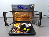 Front view of Kalorik MAXX Digital Air Fryer Oven with doors open, digital display lit, showing timer and temperature. Accessories and cookbook are visible in front of the oven.