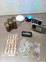 Overview of all items in the lot including three brown pillar candles, three digital clocks, one speaker with light, three packs of white LED rope lights, and one multi-color LED party light.