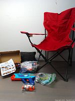 Red camping chair with various outdoor toys grouped on the floor including two water blasters, blow-up mattress in plastic, and 3D pen items with instructions.