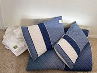 Stack of folded white bamboo sheets and bed skirt beside six blue and white chevron patterned pillows on the floor.