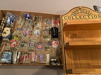 Collection of assorted shot glasses with various sports team logos including Detroit Tigers, Cincinnati Reds, Oakland A's, Phillies, and more, placed in a box.