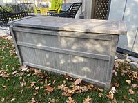 Full view of plastic deck box showing weathered gray exterior and lid, positioned on grass with fallen leaves.