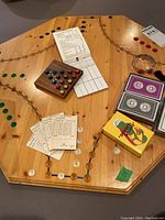 Overhead view of wooden game board with marbles in holder and multiple decks of cards, score sheets, and magnifying glass