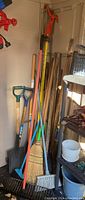 Overall view of garden tools including shovels, brooms, rakes, and long handled tools in a corner.