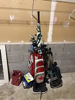 Full view of red and white golf bag holding several golf clubs, golf umbrella strapped to bag, black Sketchers golf shoes in front, a golf caddy on the right, box of golf balls on the floor beside bag.