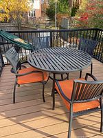 Round metal patio table with four matching chairs featuring orange cushions on a wood deck, outdoor setting with railing