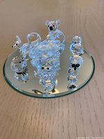 Seven clear faceted crystal animal figurines arranged on a round mirror tray. Includes swan, koala, birds, snowman, fox. Minor damage visible on some items.