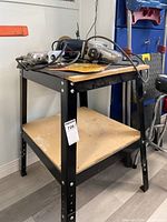 Black metal utility stand with pressboard top shelf holding some tools, showing adjustable leg holes and the lower pressboard shelf.