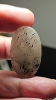 Close-up image of a hand holding an oval cabochon polished quartz gemstone with black rutilated inclusions and black tourmaline needles inside, against a dark background