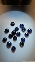 Photo showing multiple small round blue sapphire cabochons spread out on a white surface under direct light. Shows the stones' dome shape and deep blue color with visible inclusions.